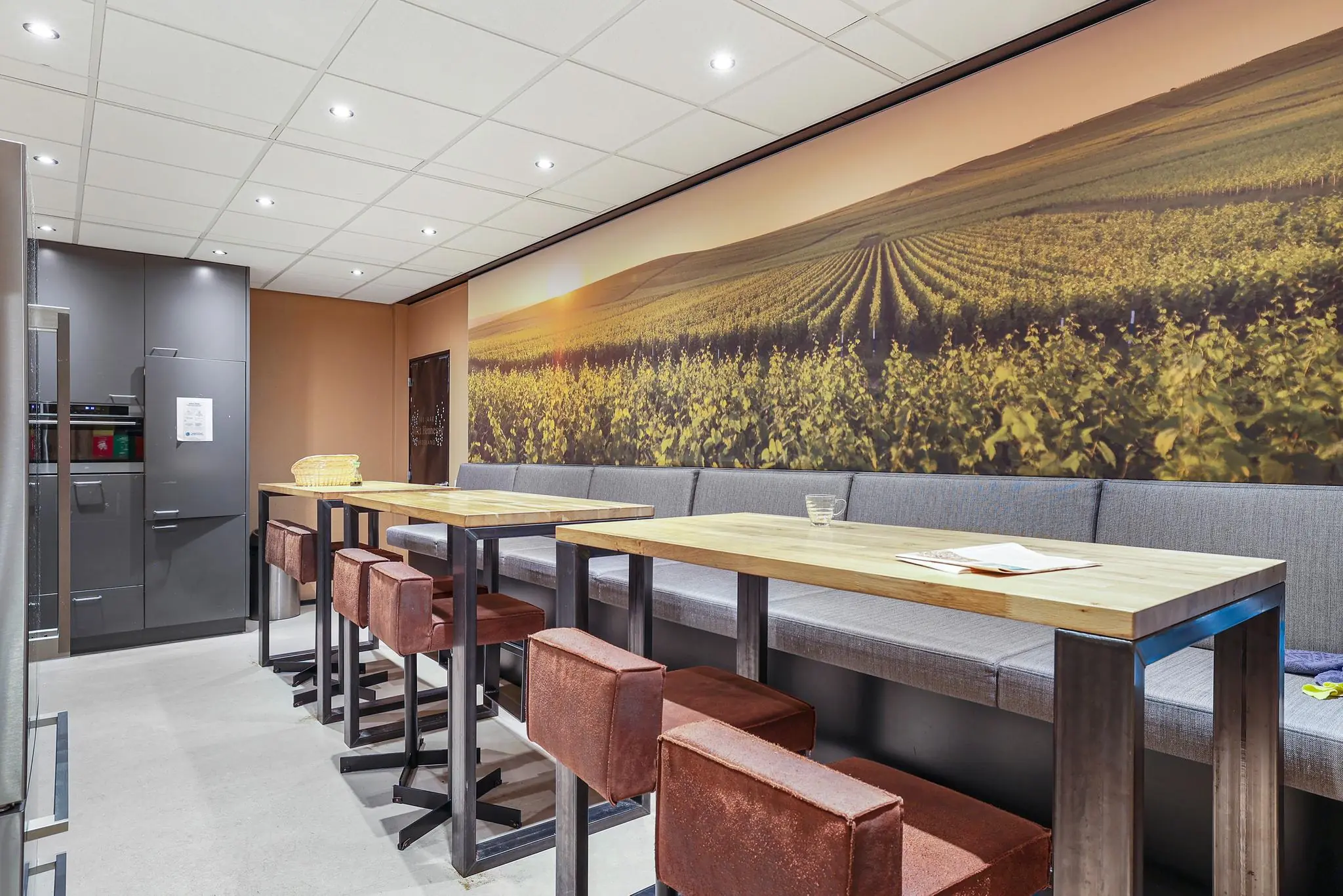 Moderne kantineruimte met hoge tafels, barkrukken en een grote wandfoto van een zonovergoten wijngaard.